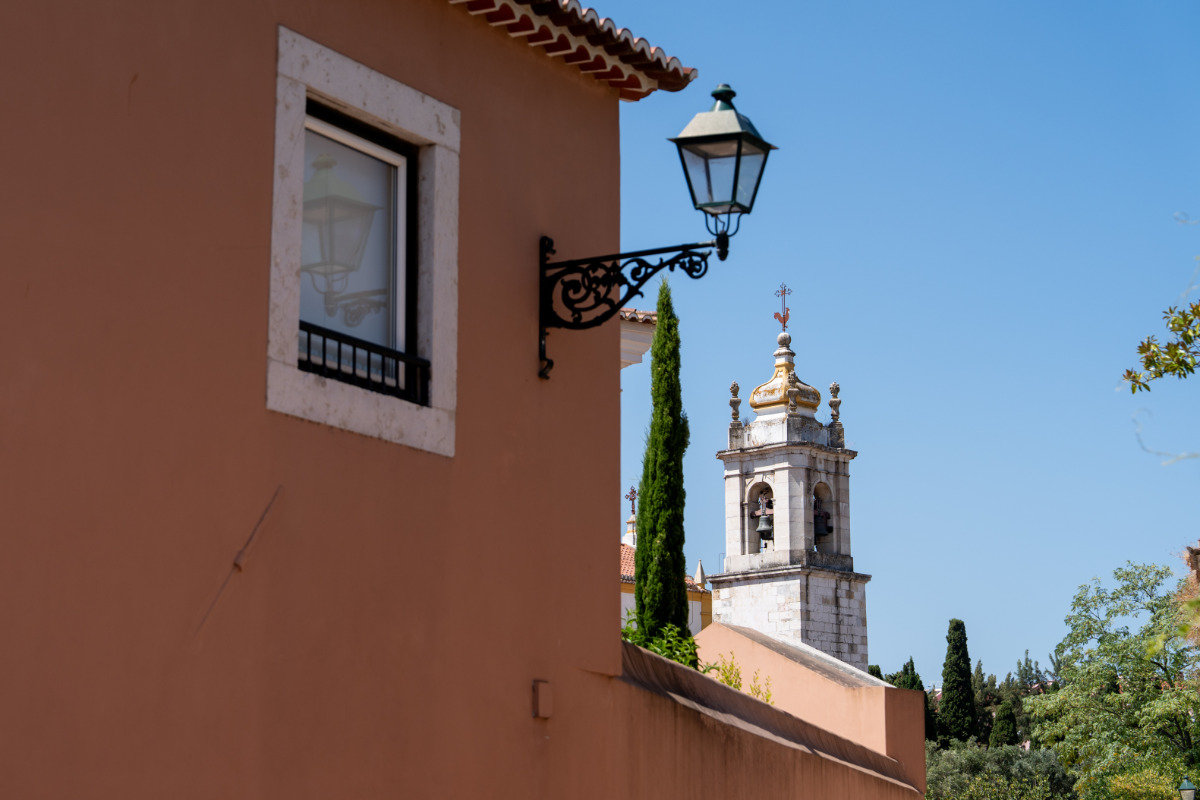 Imagem de uma rua do Lumiar, com um candeeiro de rua tradicional, uma parede de tom suave e a torre da Igreja de São João Baptista ao fundo, sob um céu azul claro.