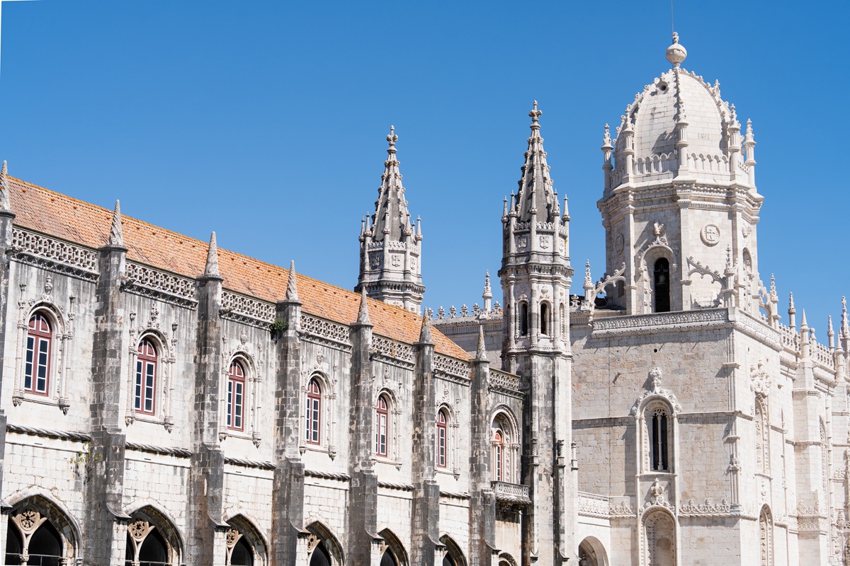 Vista detalhada do Mosteiro dos Jerónimos com arquitetura ornamentada, com torres e uma cúpula sob um céu azul claro.