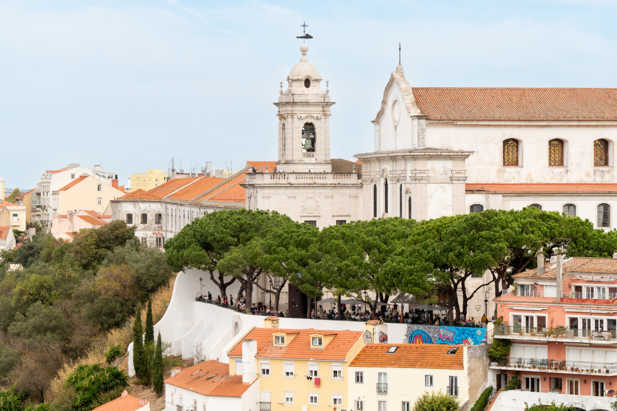 Vista panorâmica do Miradouro, Convento e Igreja da Graça. Em primeiro plano inclui árvores exuberantes e edifícios coloridos.