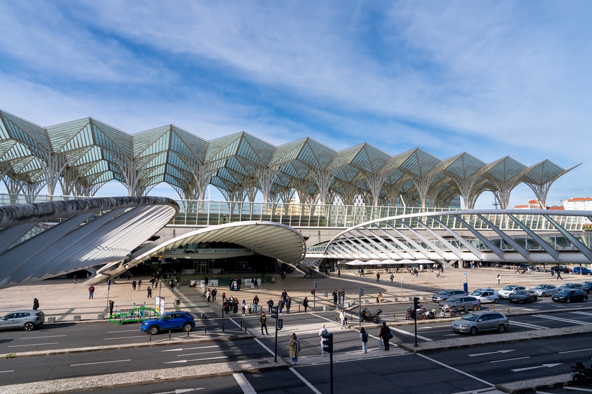 Vista panorâmica da Gare do Oriente, com telhado ondulado e arcos de vidro sob um céu azul. Pessoas, carros e bicicletas preenchem a praça movimentada, transmitindo o agitado ambiente urbano.