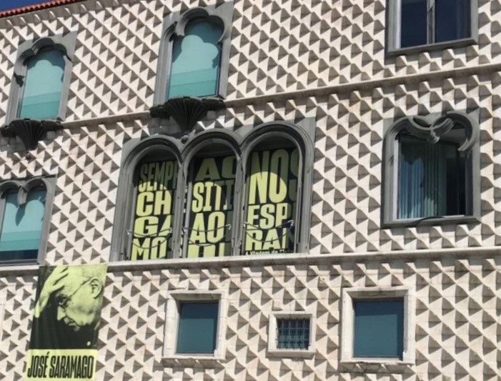 A imagem mostra a fachada geométrica da Casa dos Bicos - edifício branco com texturas piramidais e janelas em arco. Uma faixa com a imagem de José Saramago está em destaque.
