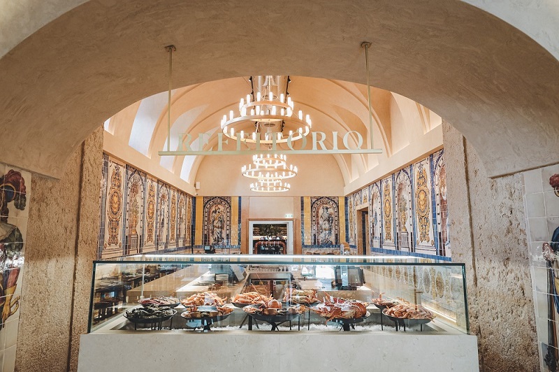 Interior da Cervejaria Trindade, no Chiado, com balcão de marisco em vidro, painéis de azulejos nas paredes e iluminação suspensa sob arcos de pedra.