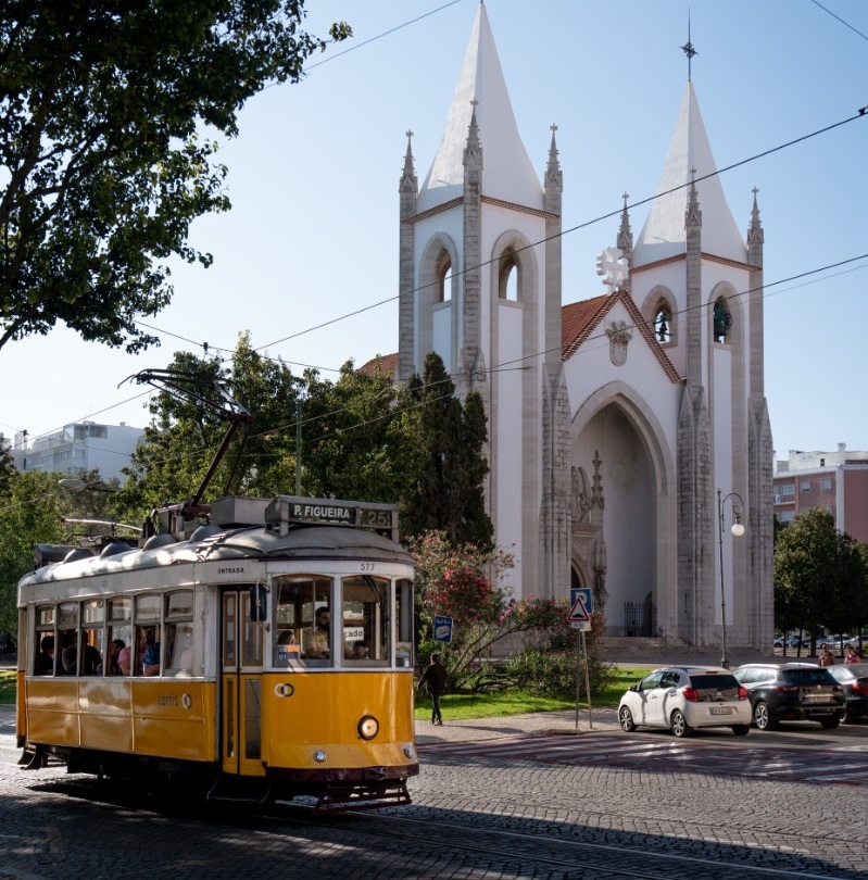 Um elétrico a passar pela igreja de Campo de Ourique, branca com torres altas, rodeada por vegetação e carros estacionados num dia ensolarado