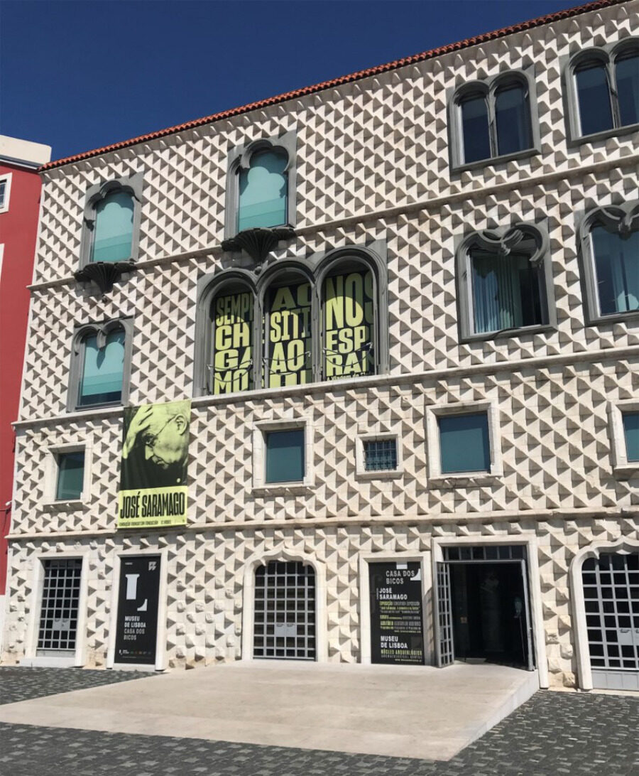 A imagem mostra a fachada geométrica da Casa dos Bicos - edifício branco com texturas piramidais e janelas em arco. Uma faixa com a imagem de José Saramago está em destaque.