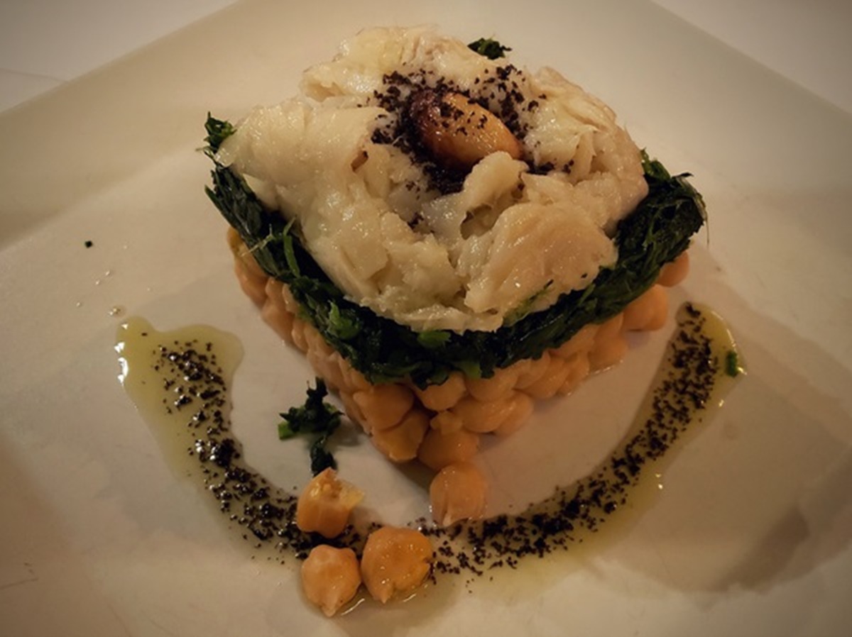 Um prato quadrado com camadas de grão-de-bico, couve e bacalhau lascado. É guarnecido com azeite e ervas, criando uma apresentação gourmet.