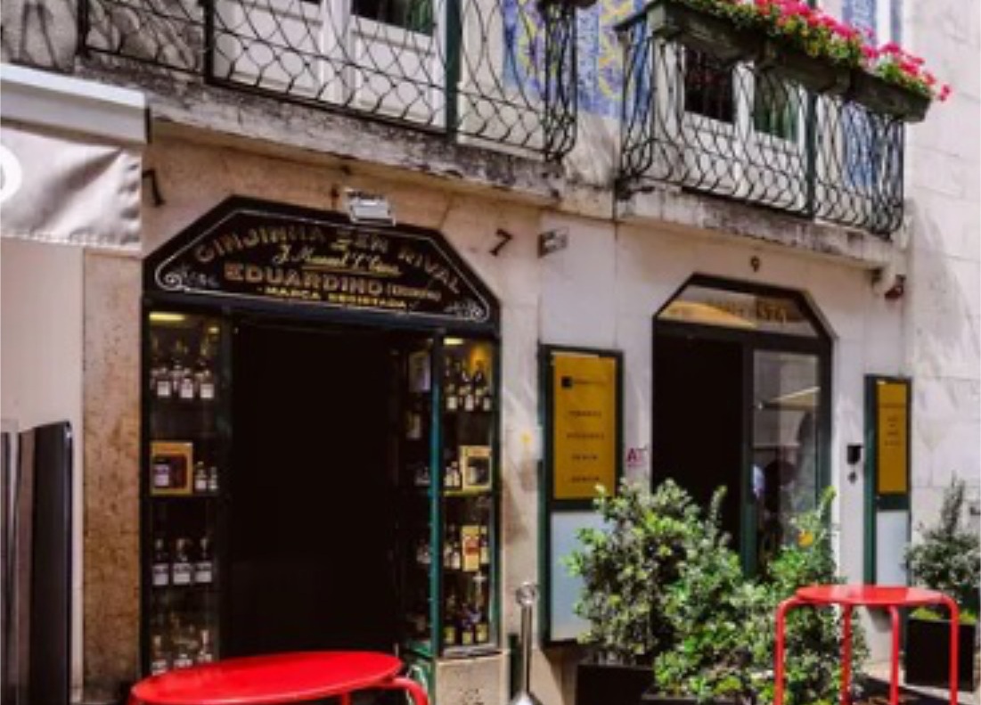 Vista da rua de um dos pequenos bares históricos na Baixa de Lisboa, com plantas em vasos e uma mesa vermelha. Uma varanda superior com flores adiciona uma atmosfera acolhedora e convidativa.