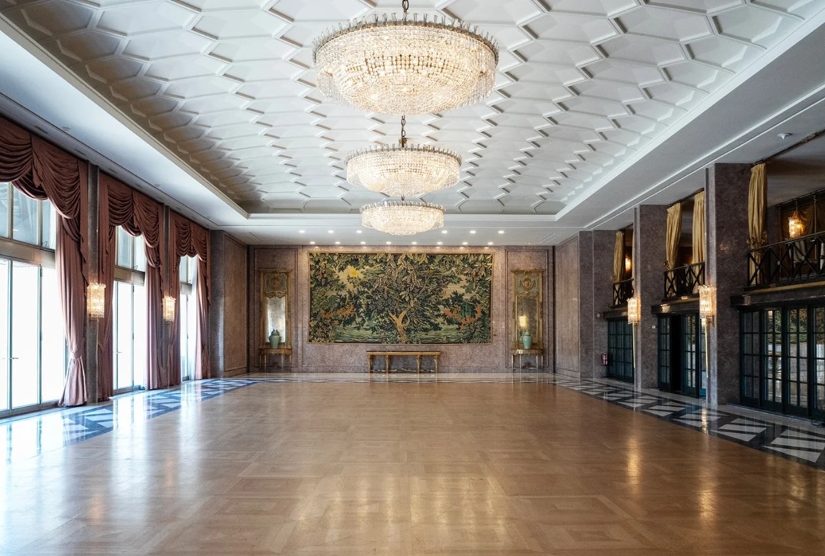 Salão de baile do Hotel Ritz, elegante, com pisos de madeira, grandes lustres e um tapete decorativo na parede. A luz do sol entra através de janelas altas com cortinas vermelhas. Ambiente luxuoso e espaçoso.