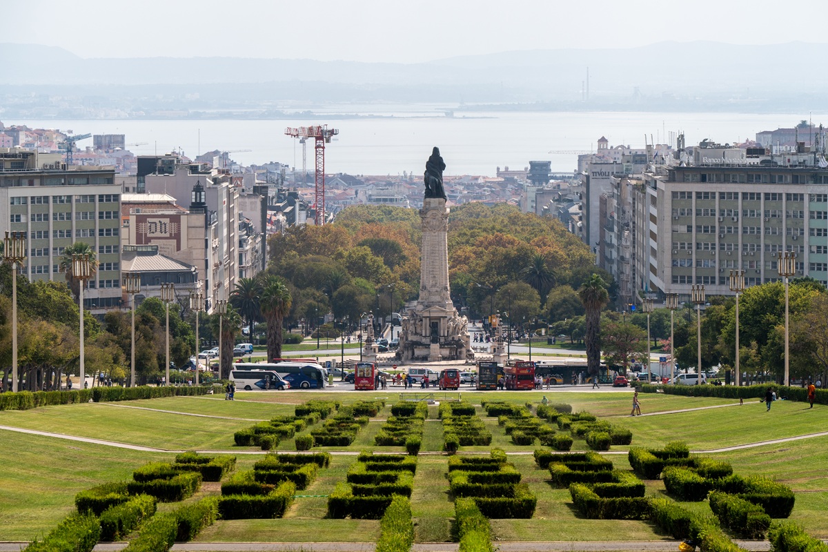Parque Eduardo VII e monumento na rotunda Marquês de Pombal, rodeada de edifícios altos, árvores e áreas verdes bem cuidadas, com o rio Tejo ao fundo.