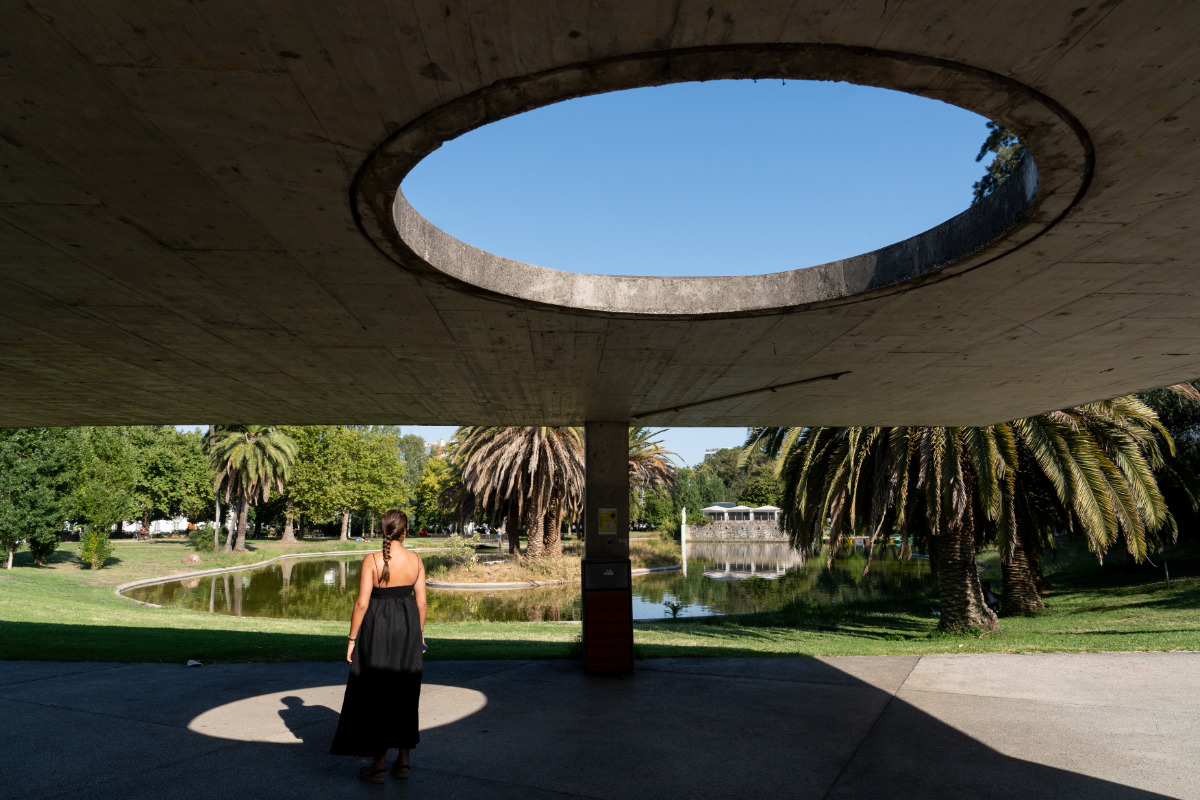 Uma jovem debaixo de uma grande estrutura de betão com uma abertura circular, olhando para o lago e árvores do Jardim do Campo Grande