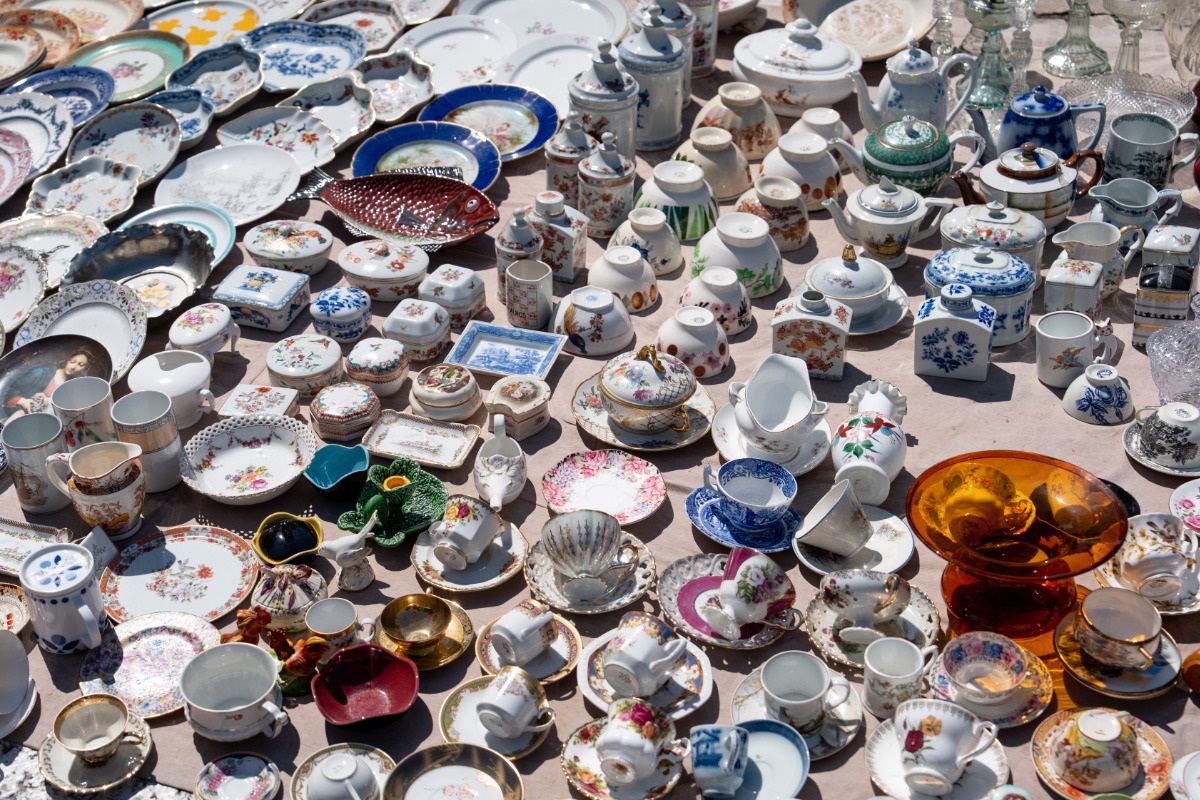 Vários pratos, xícaras e potes de porcelana de diferentes tamanhos e cores, expostos no chão para venda, na Feira da Ladra.