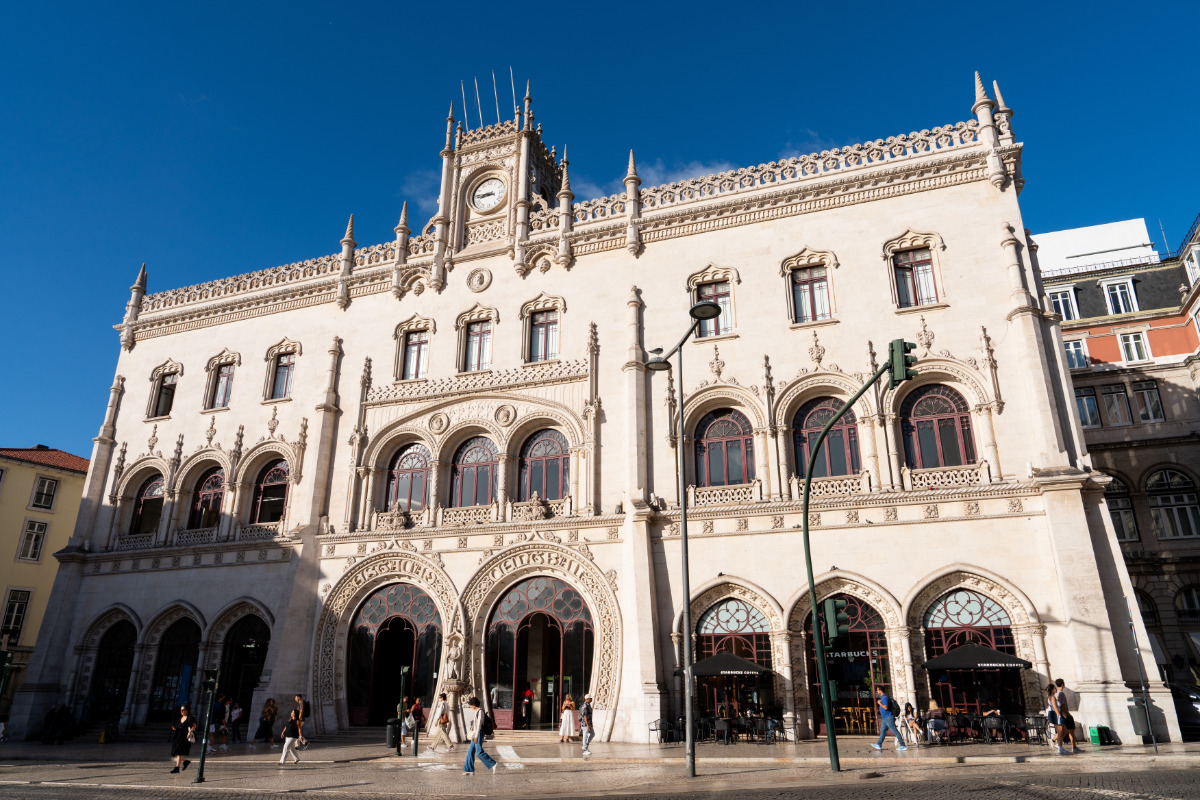 A arquitetura neomanuelina da Estação Ferroviária do Rossio em Lisboa apresenta arcos intrincados, detalhes ornamentados e um relógio central sob um céu azul claro.