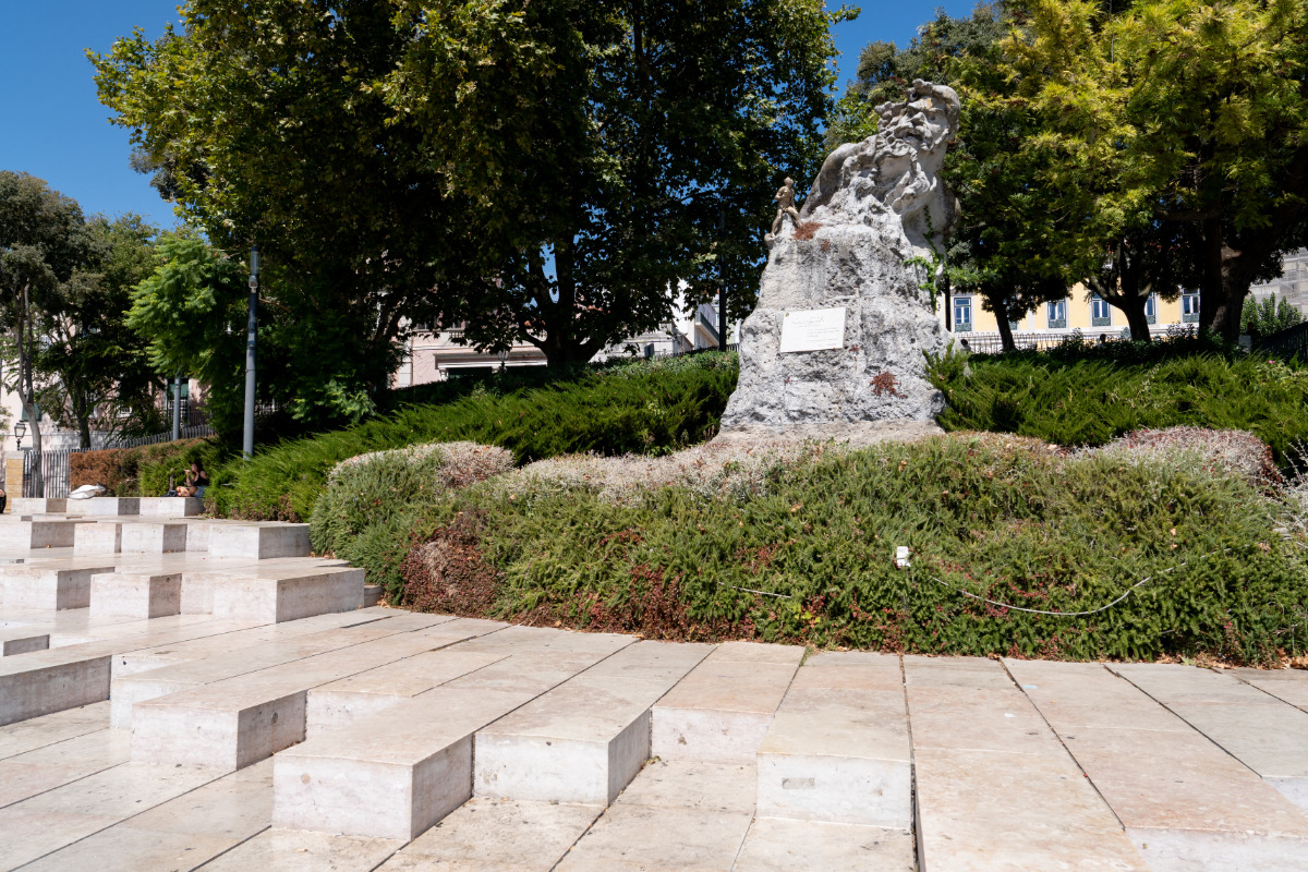 Estátua "Adamastor" em fundo de árvores verdes. À frente, piso com degraus em pedra que servem de bancos, onde os visitantes podem sentar e contemplar a cidade.