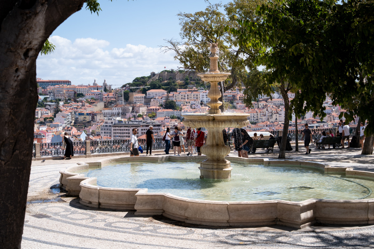Miradouro de São Pedro de Alcântara com o chafariz ao centro e várias pessoas no local. Ao fundo, avista-se a colina do castelo.