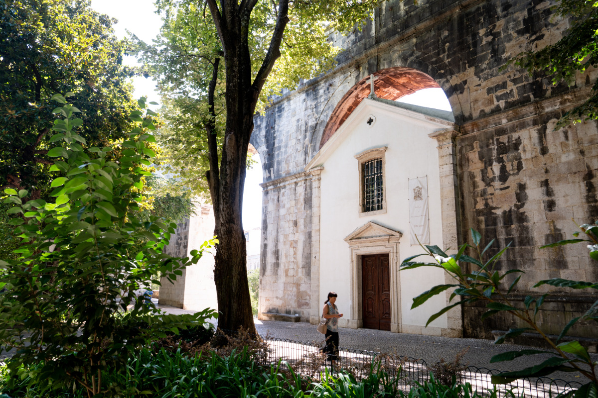 Uma pessoa passa pela pequena Capela de Nossa Senhora de Monserrate - edifício branco com uma porta de madeira, debaixo do Aqueduto das Águas Livres, rodeado por árvores e vegetação.