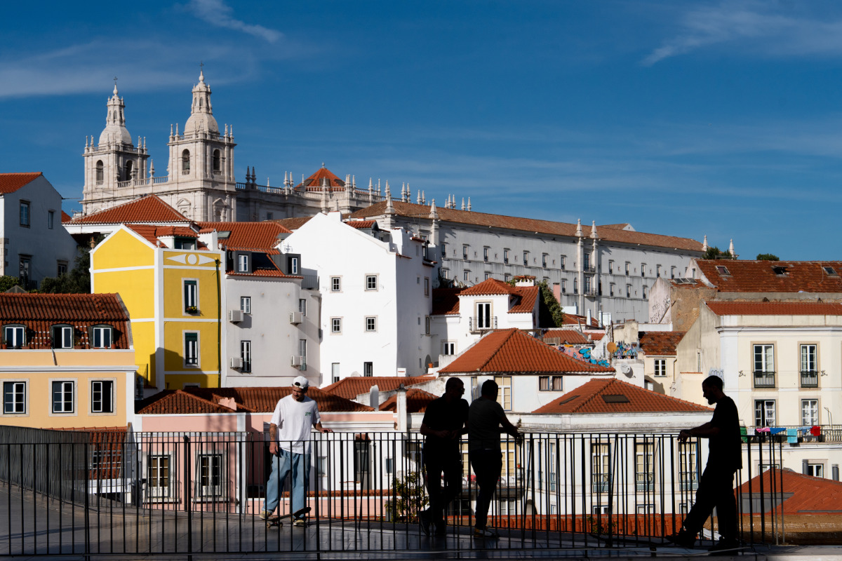 Vista de Lisboa a partir do Miradouro das Portas do Sol, com prédios históricos e pessoas caminhando. Destaca-se a arquitetura tradicional com céu azul e clima ensolarado.
