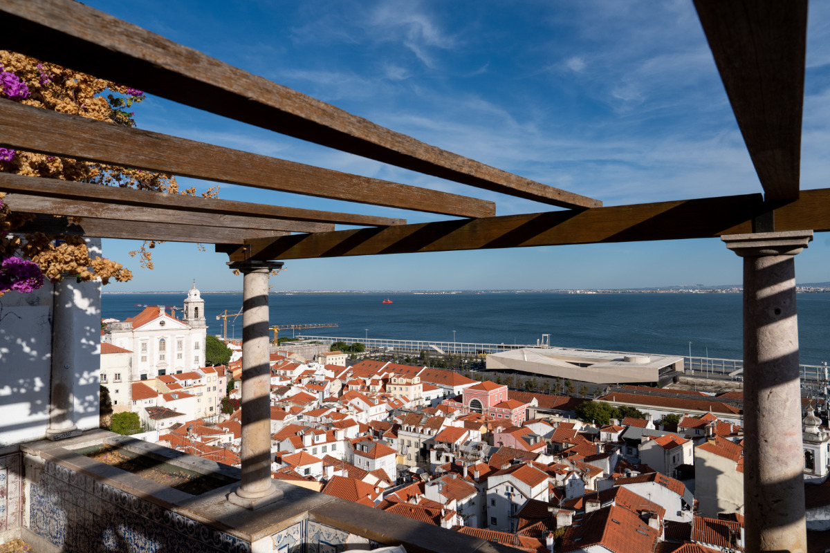 Vista panorâmica de Lisboa a partir do Miradouro de Santa Luzia, com destaque para o rio Tejo ao fundo, telhados de tijolos vermelhos, uma igreja histórica, num dia de céu claro.