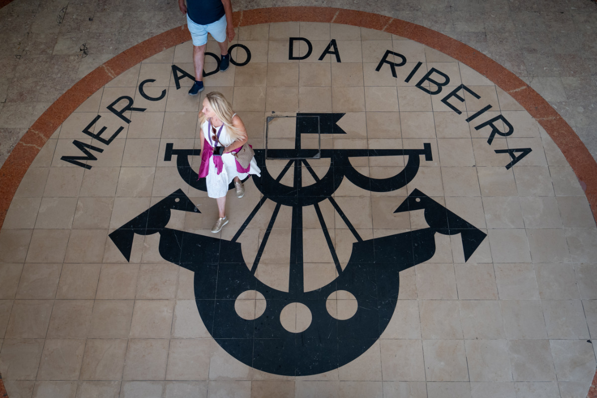 Imagem de uma mulher caminhando no centro do Mercado da Ribeira, com o logotipo do mercado no chão.