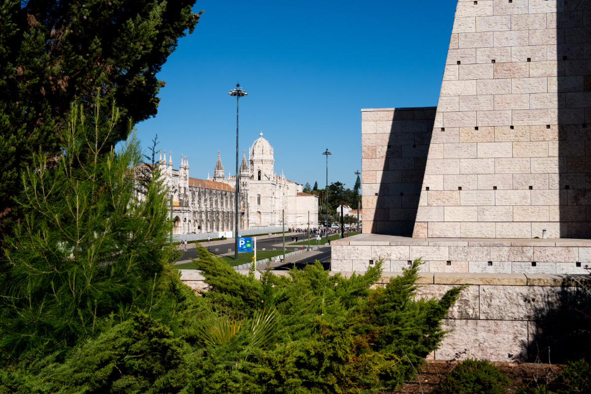 Mosteiro dos Jerónimos, monumento histórico com torres pontiagudas, emoldurado por uma vegetação exuberante e arquitetura moderna do Centro Cultural de Belém, sob um céu azul claro.