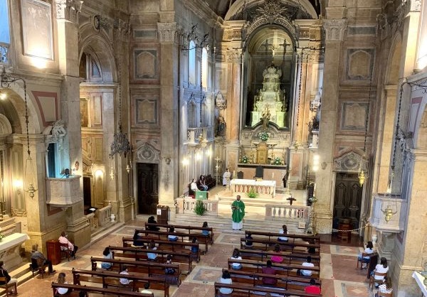 Interior da Igreja de Nossa Senhora do Amparo com altares decorativos, pilares de mármore e bancos de madeira com pessoas sentadas e padre, com vestimenta verde, no altar.