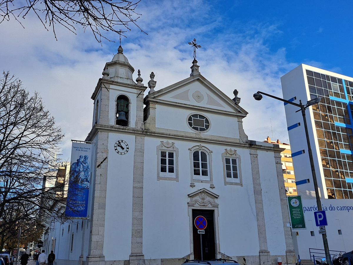 Igreja dos Santos Reis Magos, situada no Campo Grande, com arquitetura clássica, vista frontal, detalhes das janelas e relógio, céu azul ao fundo, ao lado de edifícios modernos, cenário urbano.