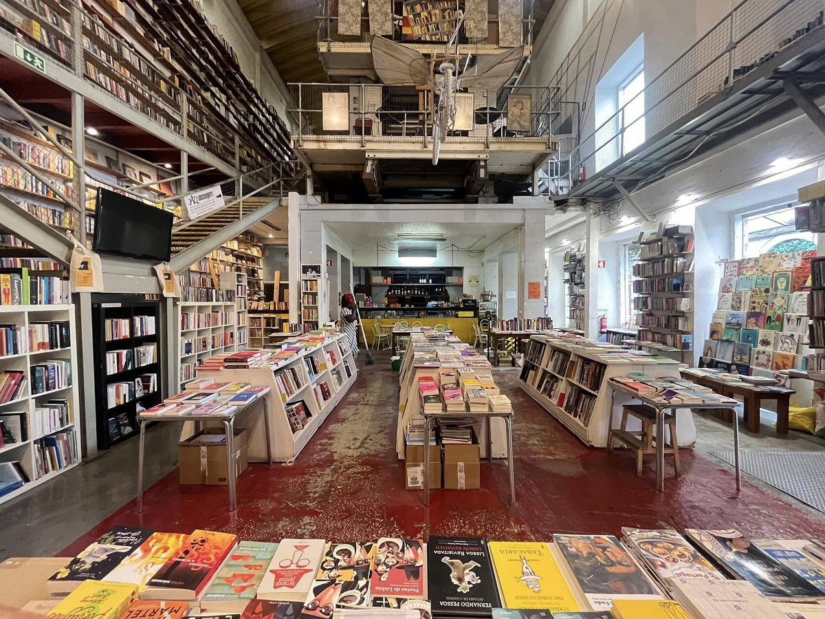 Imagem da livraria Ler Devagar com várias prateleiras cheias de livros, mesas com livros dispostos para venda e espaço amplo com iluminação natural.