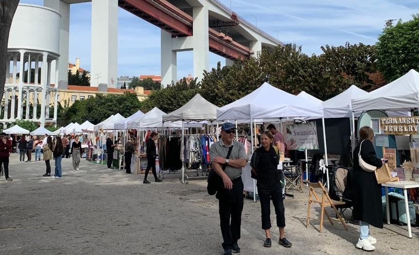 Feira ao ar livre na LX Factory, com várias tendas de venda de roupa, acessórios e objetos de decoração. Os visitantes caminham e observam o que está exposto para venda.