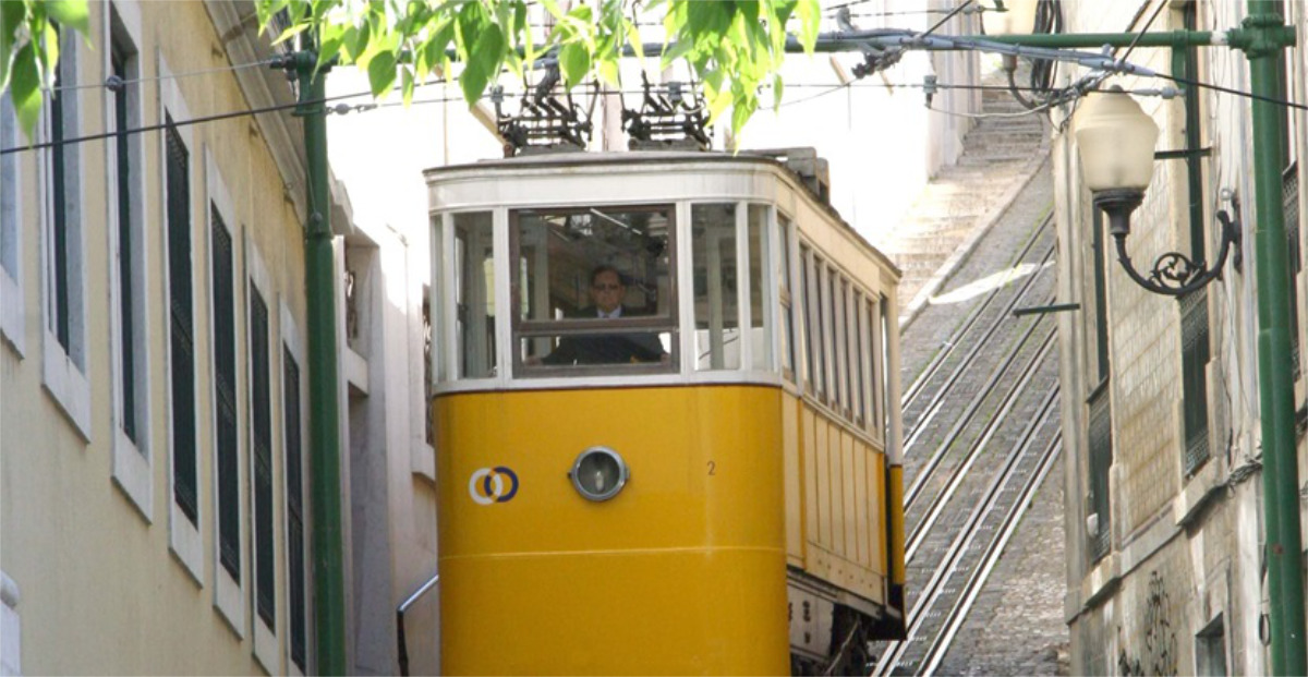 Ascensor do Lavra percorre uma rua estreita, ladeada por edifícios e trilhos ascendentes num cenário ensolarado.