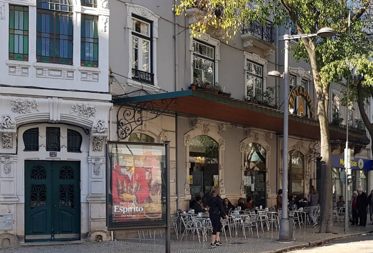 Vista do exterior ornamentado do café histórico A Tentadora. Pessoas sentadas na esplanada em mesas prateadas sob as árvores. No toldo, acima da porta de entrada, uma placa em arco onde se lê "A Tentadora".