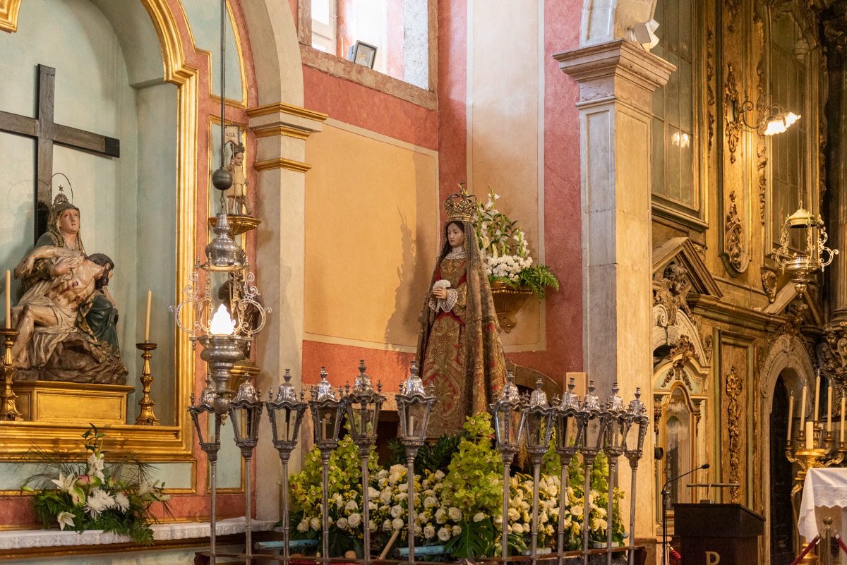 Interior da Capela de Nossa Senhora da Saúde, focando a área do altar. Em primeiro plano, há uma série de castiçais de prata ornamentados dispostos em linha, com velas acesas, emitindo um brilho quente. Diante desses castiçais, encontram‑se arranjos florais, incluindo lírios brancos e folhagem verde, acrescentando à atmosfera solene e reverente.