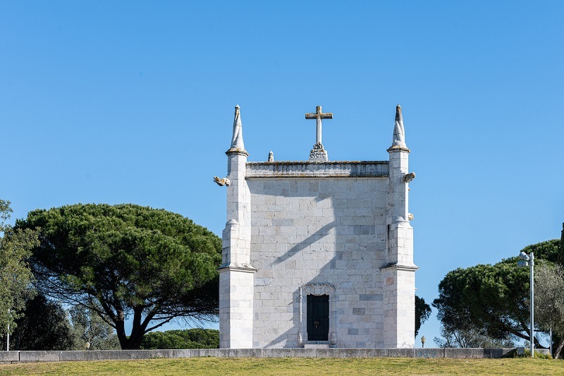 Fotografia da fachada da Capela de São Jerónimo, de pedra branca com uma cruz no topo, situada em uma área aberta cercada por vegetação sob um céu azul claro.