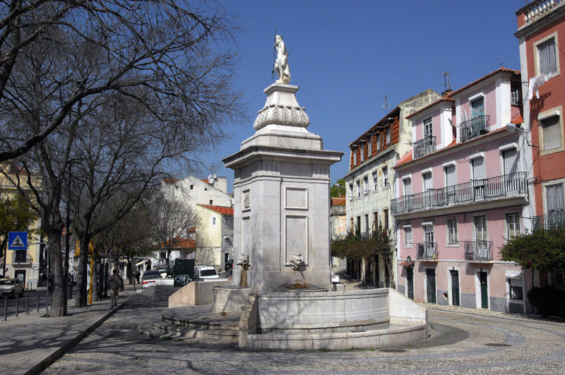 Chafariz da Praça da Armada, rodeado por árvores e edifícios coloridos sob um céu azul claro. Pessoas caminham nas proximidades.