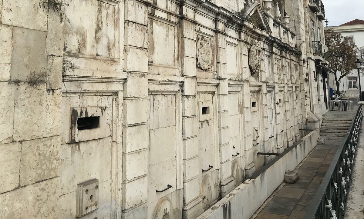 Fachada do Chafariz d’El-Rei em pedra, com relevos e inscrições desgastadas pelo tempo, e uma passarela estreita e pavimentada, paralela, delimitada por uma grade metálica.