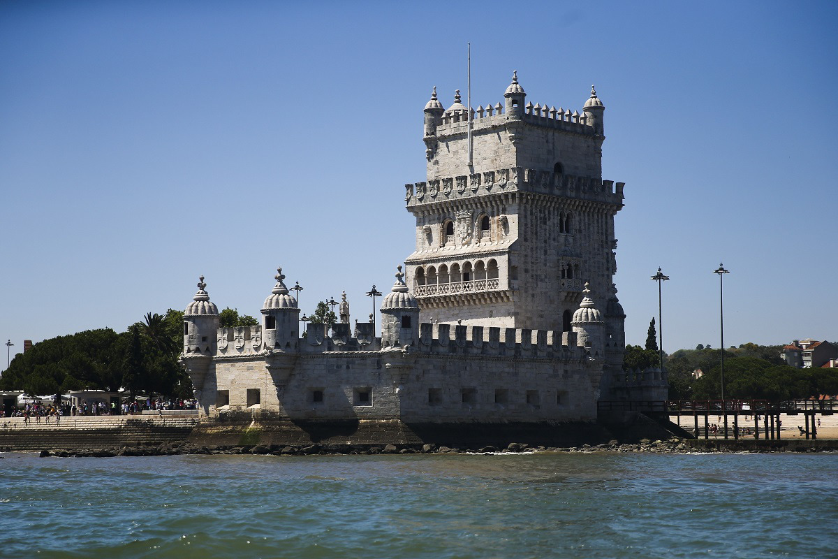 Torre de Belém, de pedra com ameias, erguida à beira-rio, rodeada por árvores e um céu azul límpido