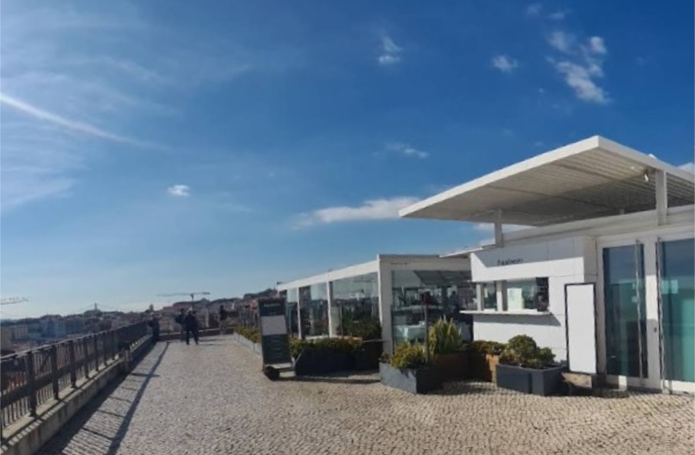 Plataforma de acesso ao Elevador, na Colina do Castelo, com um moderno café no terraço com paredes de vidro, rodeado por uma vegetação exuberante, sob um céu azul límpido, com pessoas a passear nas proximidades.