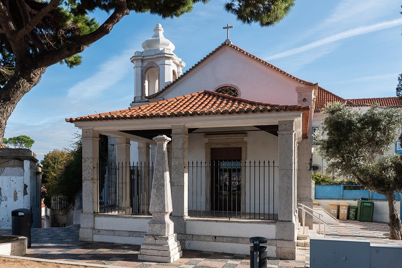 Fotografia da pequena capela branca de estilo tradicional com telhado de telhas vermelhas e uma torre de sino. A capela tem um design simples com entrada central flanqueada por duas colunas que sustentam um beiral. Uma cruz é visível no topo da torre de sino, indicando seu propósito religioso. A estrutura está cercada por uma cerca de metal, e há árvores e alguma vegetação ao fundo.
