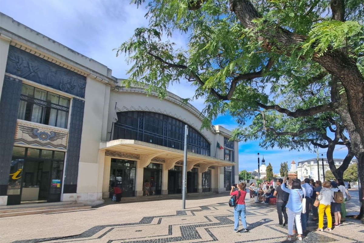Fachada da entrada principal da Estação Ferroviária do Cais do Sodré. Grupo de pessoas a apreciar e a fotografar num dia de céu azul.