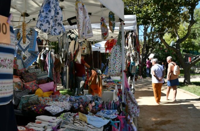 Ambiente da Feira da Luz ao ar livre com bancas de artesanato, roupas e acessórios, sob céu azul, no jardim com o mesmo nome, com árvores e visitantes passeando.