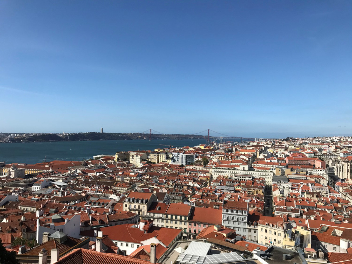Vista panorâmica de Lisboa a partir do Miradouro do Castelo de São Jorge, com o rio Tejo ao fundo e a ponte 25 de Abril em destaque sob céu claro, mostrando uma cidade de telhados vermelhos.