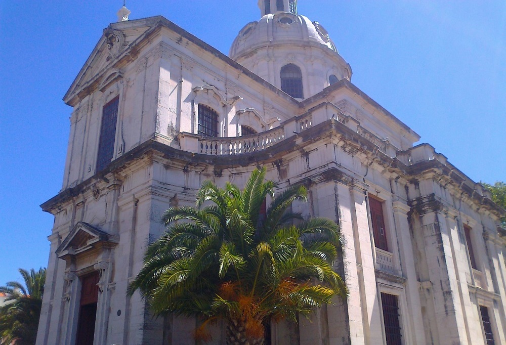 Fotografia da Igreja da Memória, que combina arquitetura barroca e neoclássica. A estrutura apresenta uma cúpula proeminente com uma cúpula menor sobre ela, adornada com detalhes intrincados e elementos decorativos. A fachada é feita de pedra de cor clara e inclui grandes janelas e portas arqueadas, algumas das quais têm portas de madeira.
