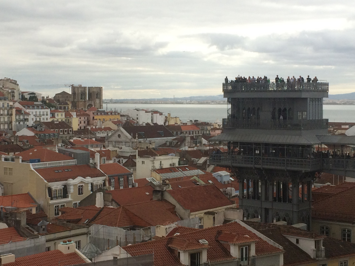 Vista dos telhados de Lisboa com o Elevador de Santa Justa em destaque; céu nublado e o rio tejo ao fundo.