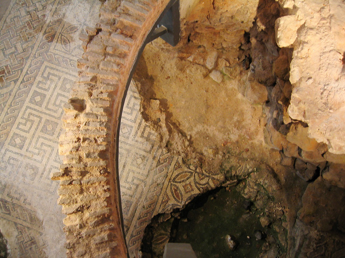 Um chão mosaico antigo parcialmente intacto com padrões geométricos está rodeado por paredes de pedra em ruínas. 