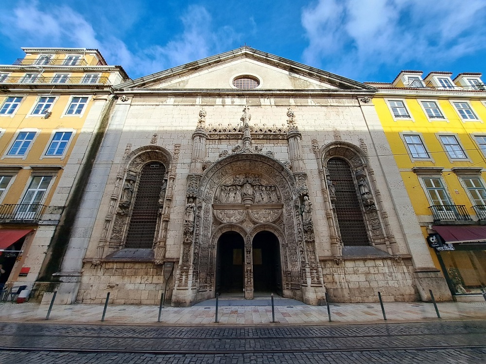 Fotografia da fachada histórica da Igreja da Conceição Velha em primeiro plano, com uma rua de paralelepípedos imediatamente à frente e uma fileira de edifícios amarelos com varandas ao fundo, sob um céu azul claro com algumas nuvens dispersas.  