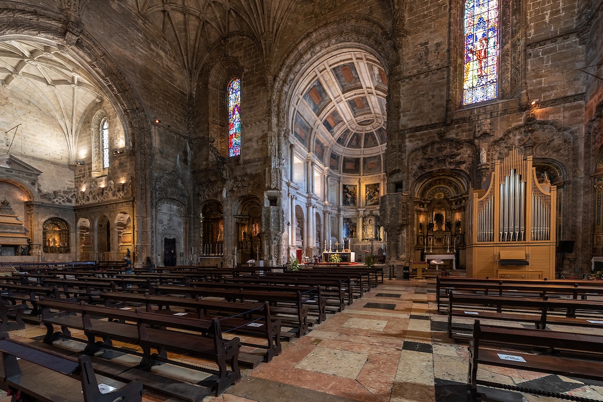 Interior da Igreja dos Jerónimos com bancos de madeira, vitrais coloridos e órgão, destacando o teto e detalhes arquitetónicos históricos.