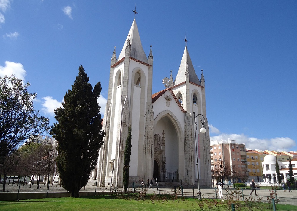 Igreja do Santo Condestável com torres altas e fachada branca, localizada em uma praça com árvores e edifícios ao fundo, sob céu azul claro.