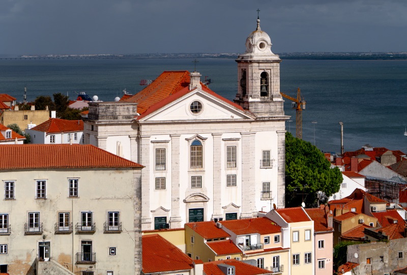 Igreja de Santo Estêvão com fachada branca e detalhes clássicos, localizada em área urbana com telhados de cerâmica vermelha, ao lado do rio Tejo, sob céu parcialmente nublado.