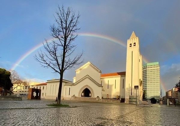Fachada da Igreja de São João de Deus com arco-íris ao fundo, céu nublado e árvore sem folhas, transmitindo paz e esperança.