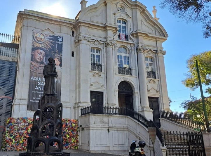 Igreja de Santo António, em estilo barroco, com fachada branca, detalhes ornamentais e uma escadaria na frente. Ao lado, escultura de Santo António e árvores verdes.