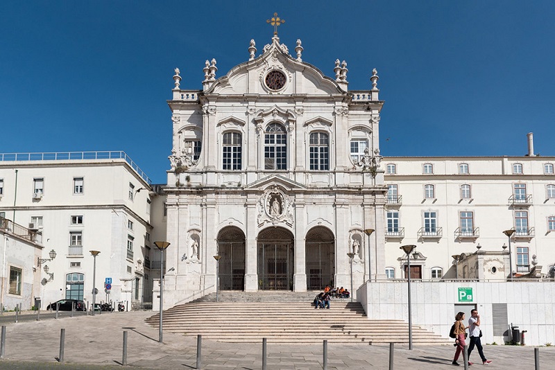 Fotografia da fachada da Igreja de Nossa Senhora das Mercês, grandiosa e ornamentada com estilo arquitetónico clássico, com pedra trabalhada intricada, grandes janelas arqueadas e uma cruz proeminente no topo. A igreja é o foco central da imagem, situada numa praça urbana.
