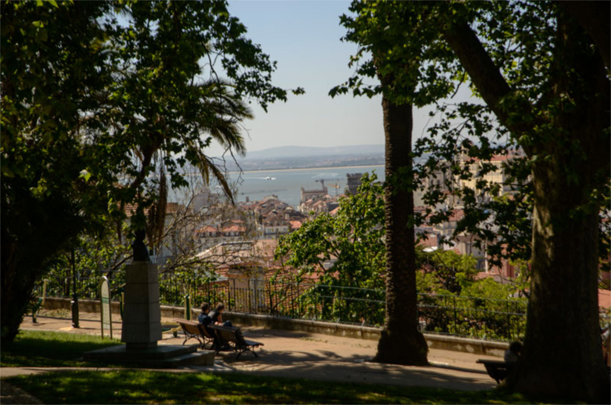 Duas pessoas sentadas num banco do Jardim do Torel, à sombra das árvores em redor, observando a cidade e o rio Tejo.