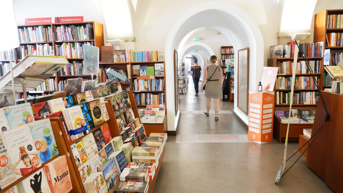 Interior da Livraria Bertrand com prateleiras cheias de livros, pessoas a caminhar pelo corredor, num ambiente acolhedor e bem iluminado.